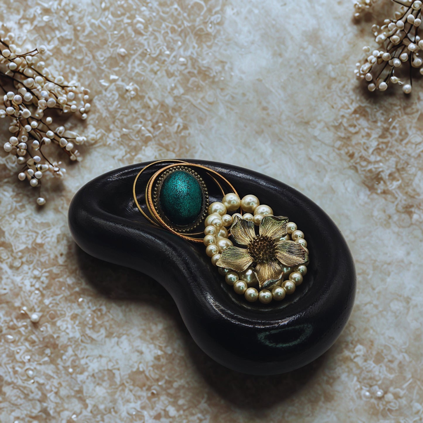 Decorative jellybean key bowl with a black base, gold and green circular design, and pearl flower on a textured beige surface.