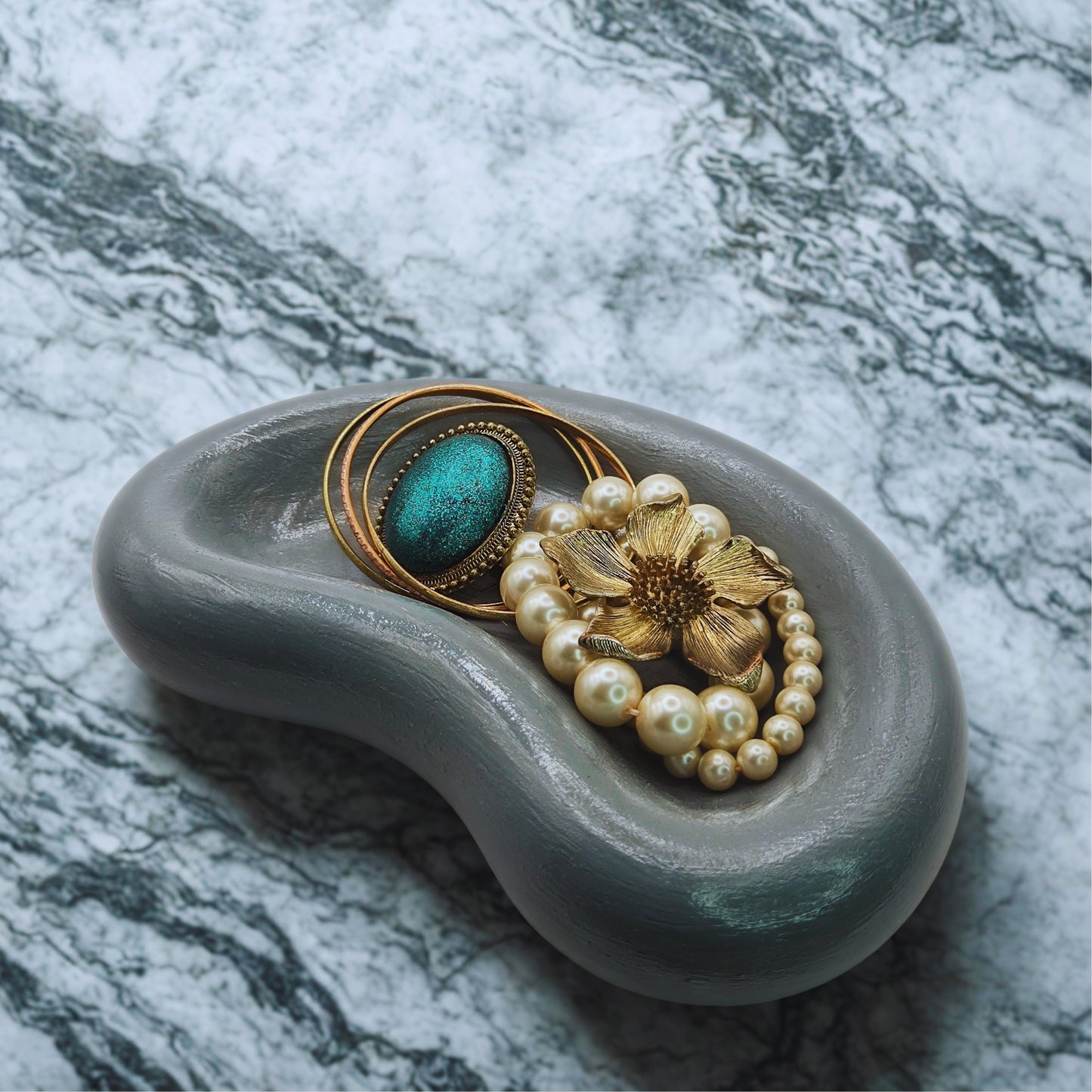 Decorative jellybean key bowl with a gold brooch, pearl necklace, and turquoise stone on a marble background