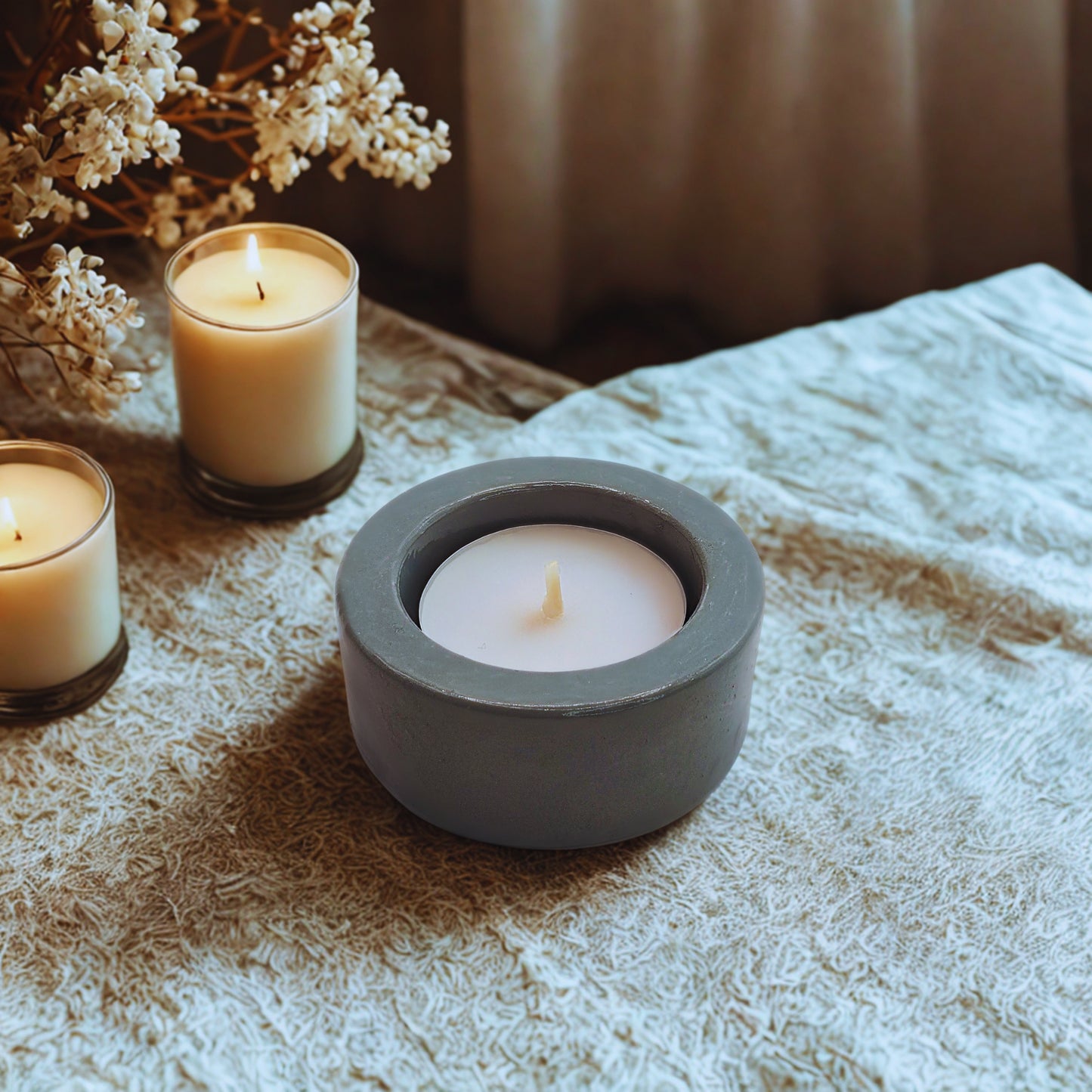 Candle in a gray holder on a textured surface with other candles and flowers in the background.
