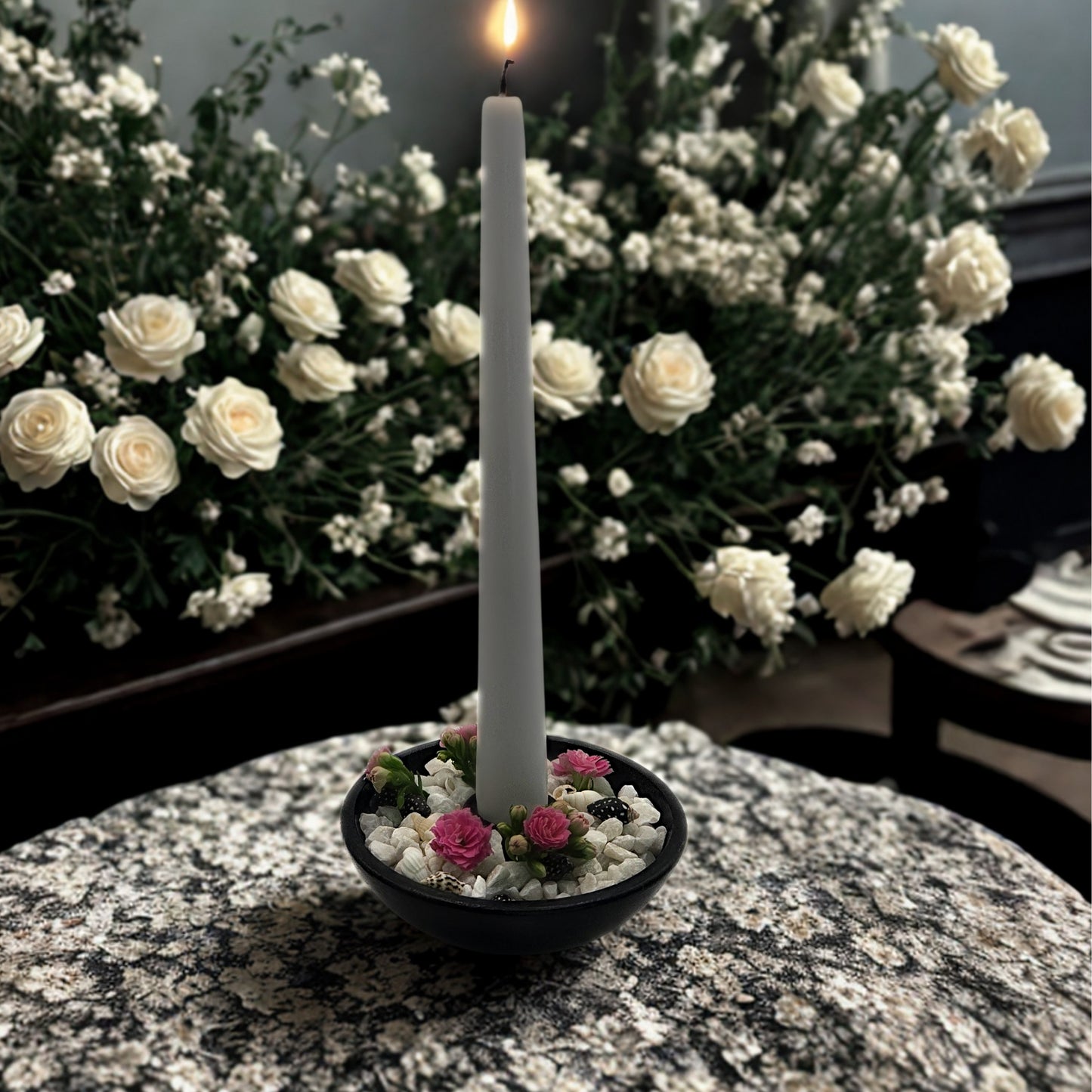 White candle in a holder with small flowers on a marble surface, surrounded by white flowers in the background.