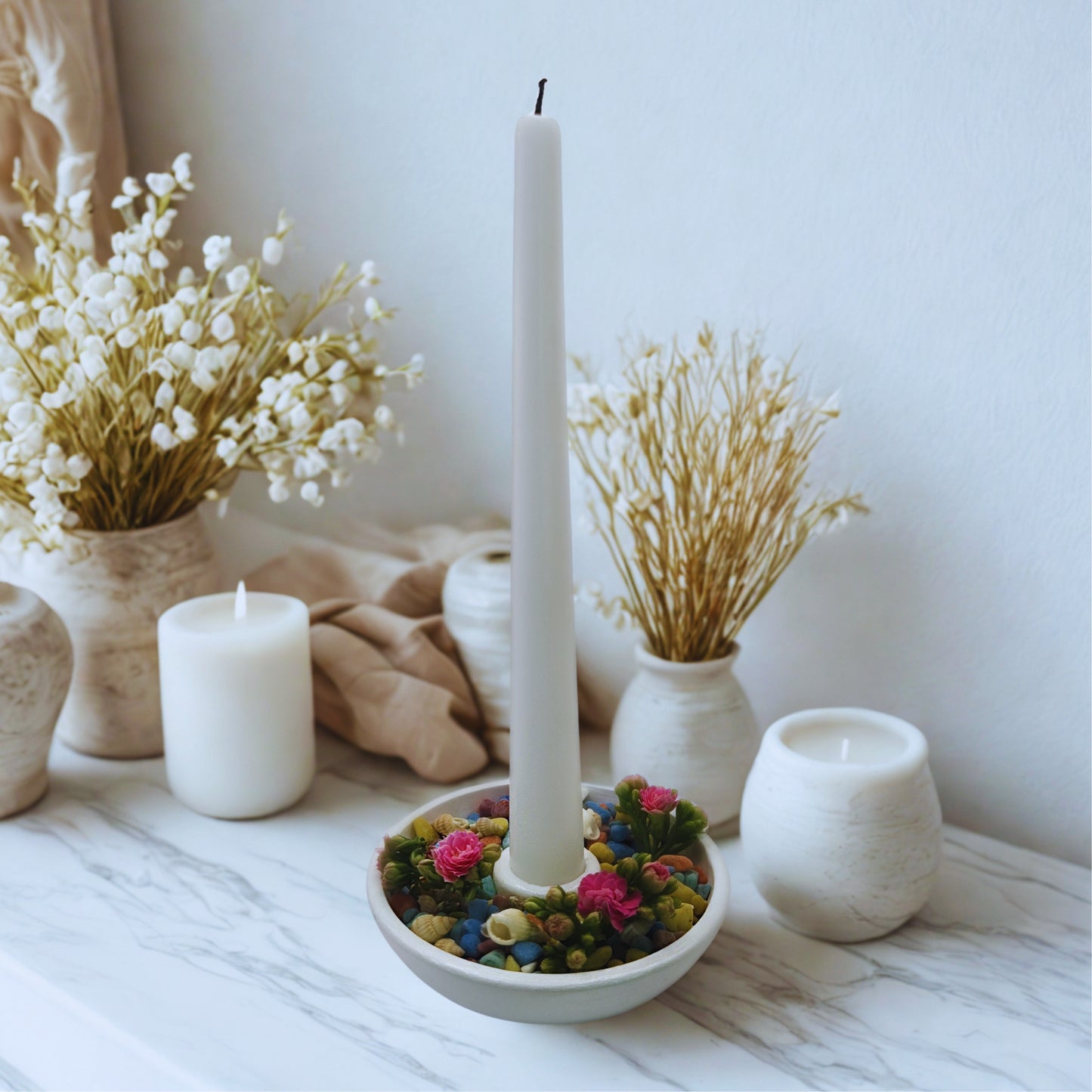 Decorative candle holder with colorful stones, surrounded by white candles and dried plants on a marble surface.