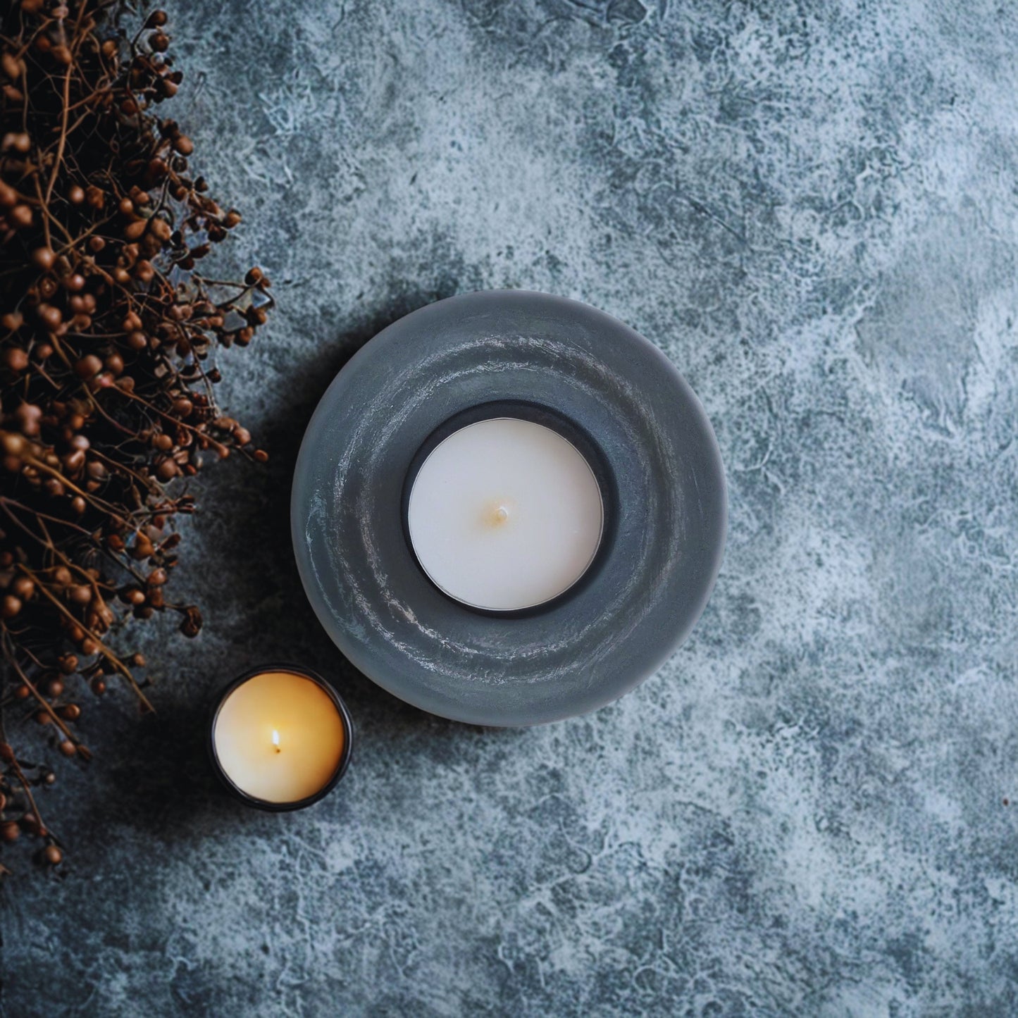 Two candles in gray holders on a textured gray surface with dried plants.