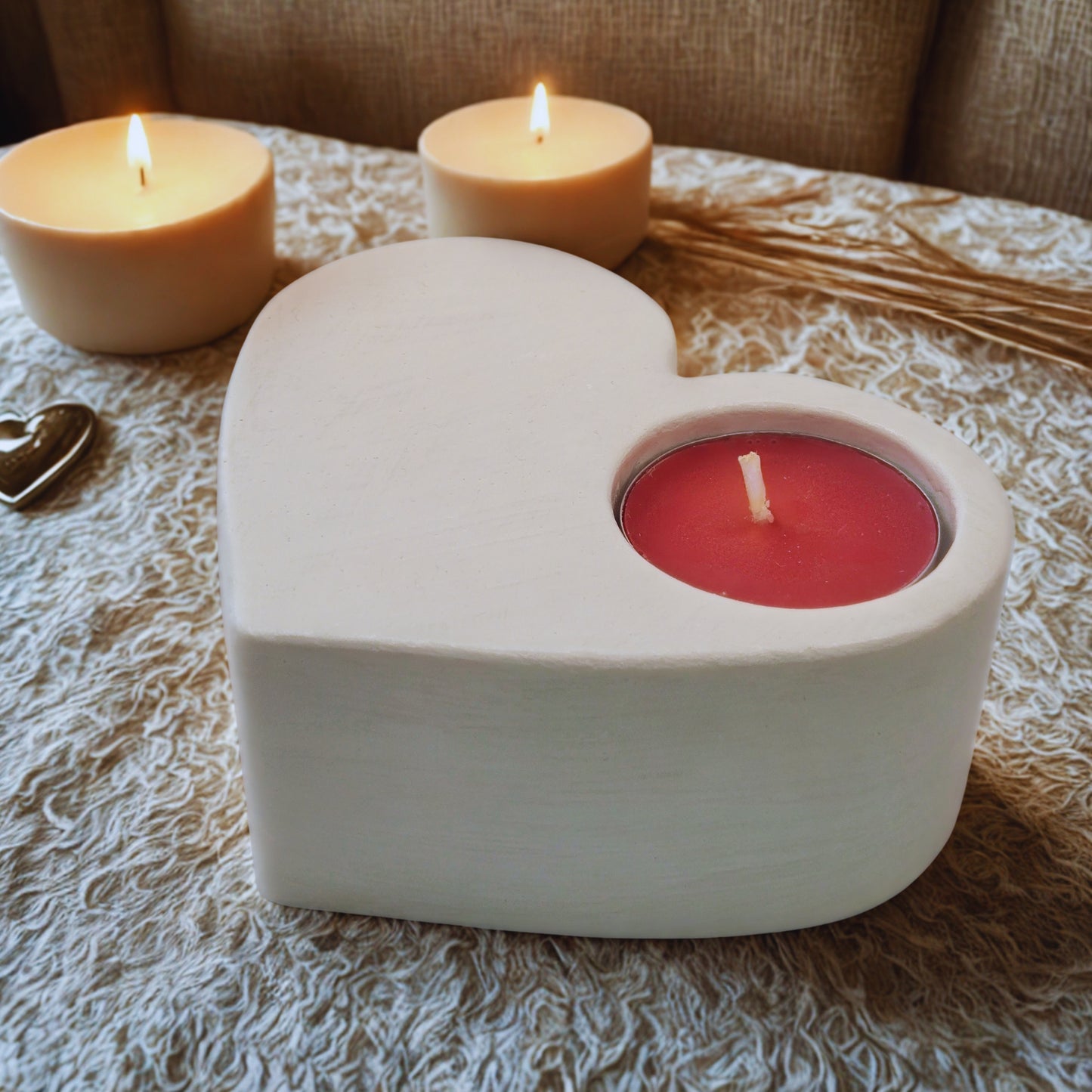 Heart-shaped candle holder with a red candle on a textured surface with two lit candles in the background.
