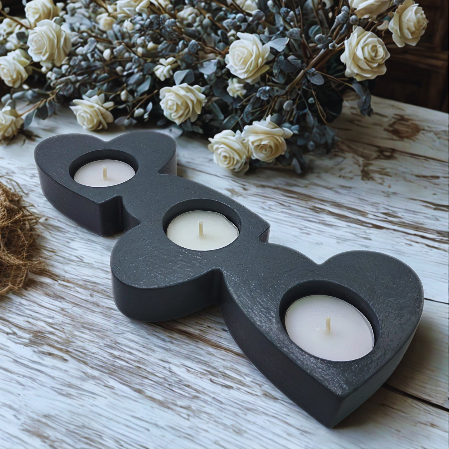 Heart-shaped candle holder with three candles on a wooden surface with flowers in the background