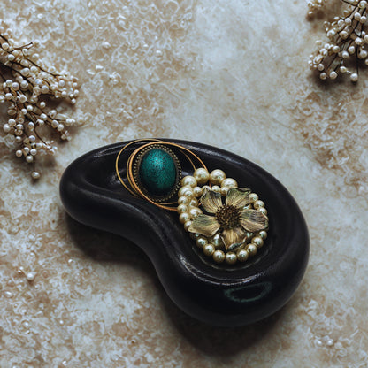 Decorative jellybean key bowl with a black base, gold and green circular design, and pearl flower on a textured beige surface.