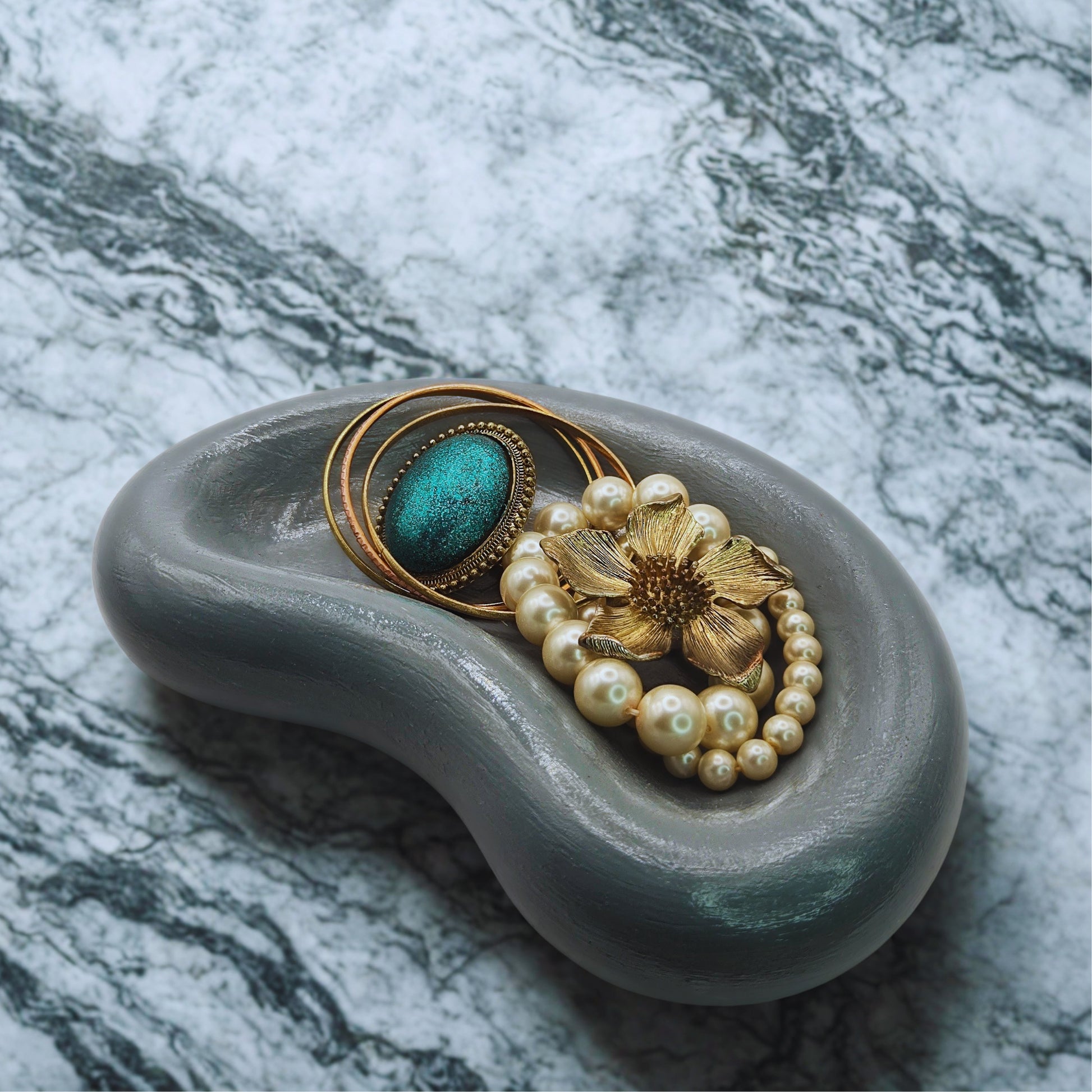 Decorative jellybean key bowl with a gold brooch, pearl necklace, and turquoise stone on a marble background