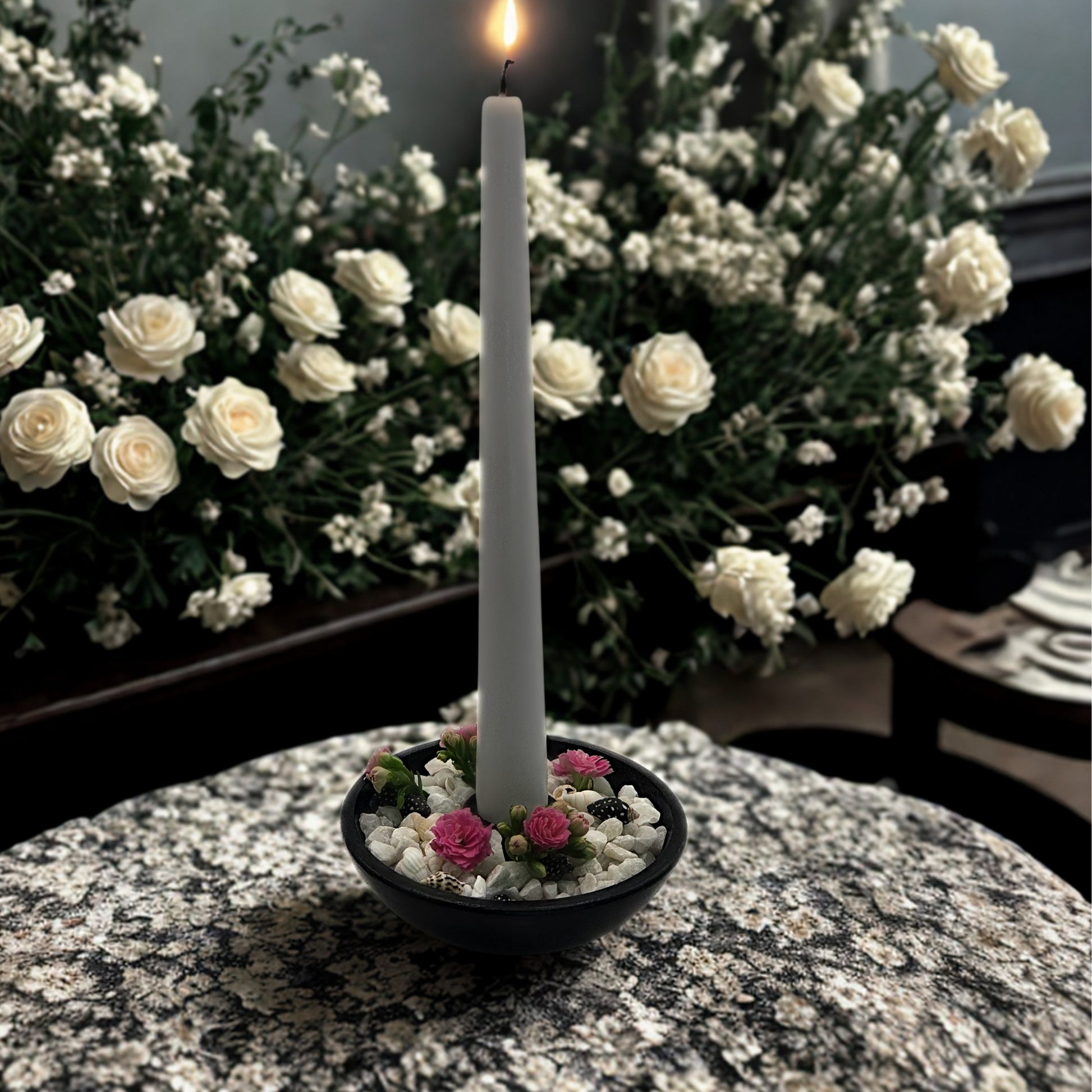 White candle in a holder with small flowers on a marble surface, surrounded by white flowers in the background.