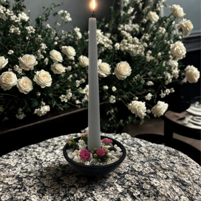 White candle in a holder with small flowers on a marble surface, surrounded by white flowers in the background.