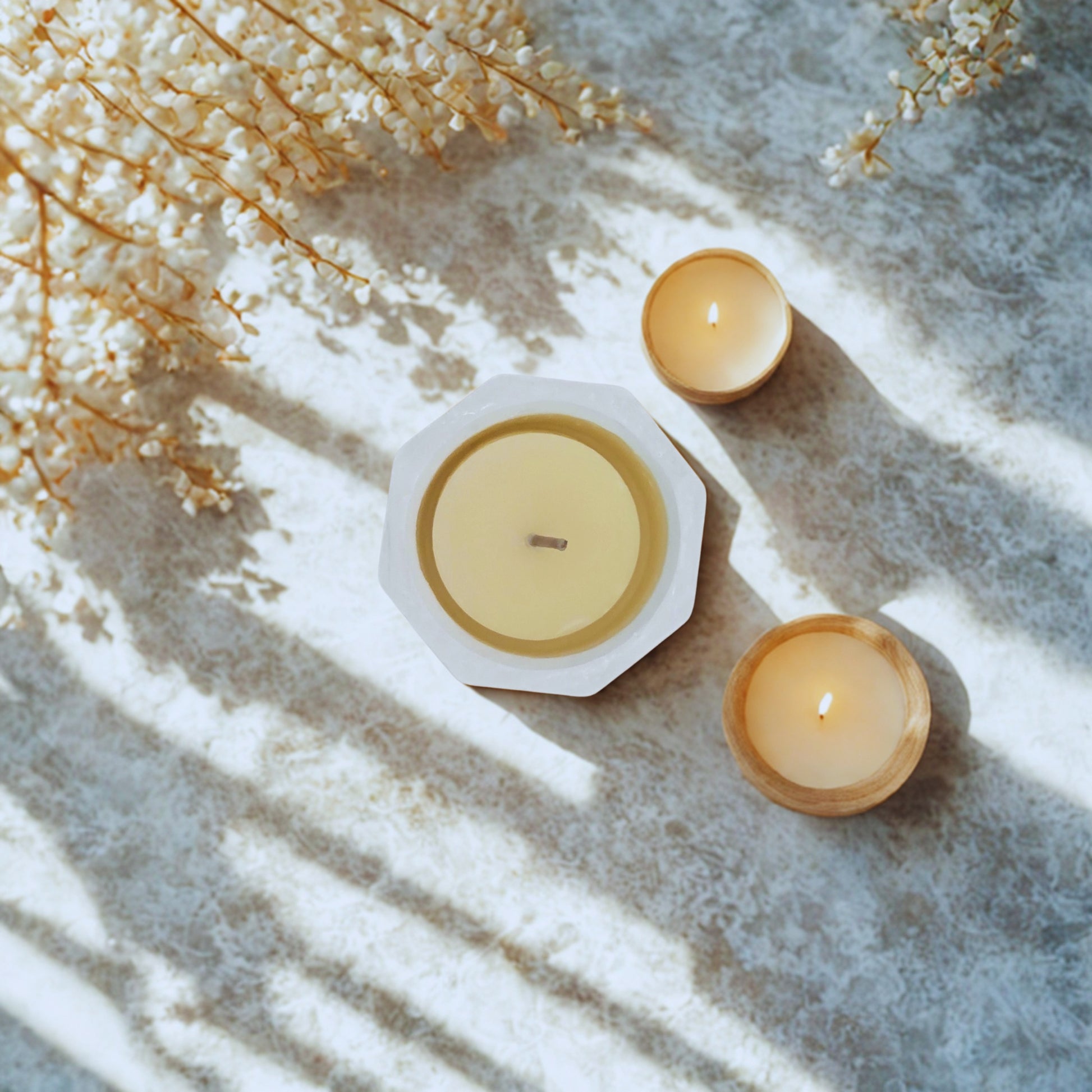 Two candles in hexagonal holders on a textured surface with dried flowers.